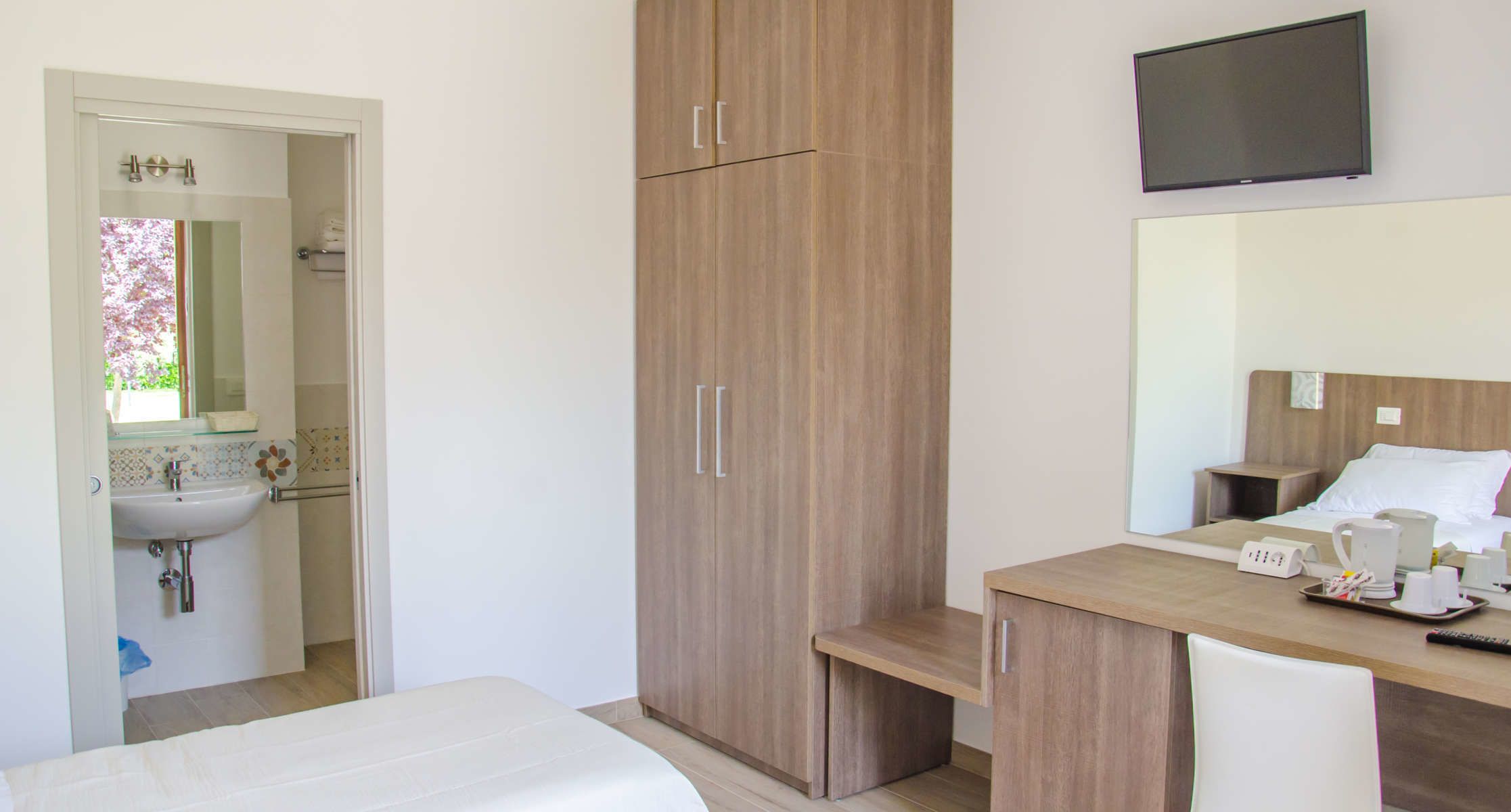 Een hotelkamer met een bed, een houten kledingkast, een nachtkastje met koffiezetapparaat, en een aan de muur bevestigde televisie.