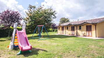 Un parque infantil con un tobogán y columpios, un edificio pequeño con dos puertas y una zona verde.