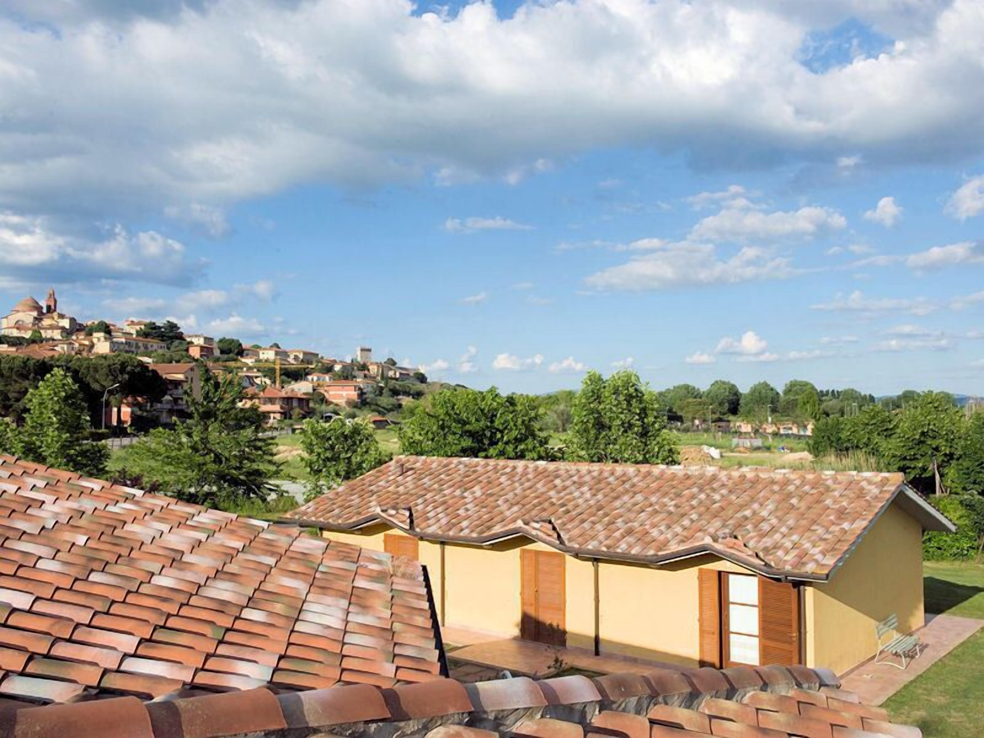 A residential area with terracotta-tiled roofs, a small building with a door and windows, and a backdrop of a hill with scattered houses and greenery.