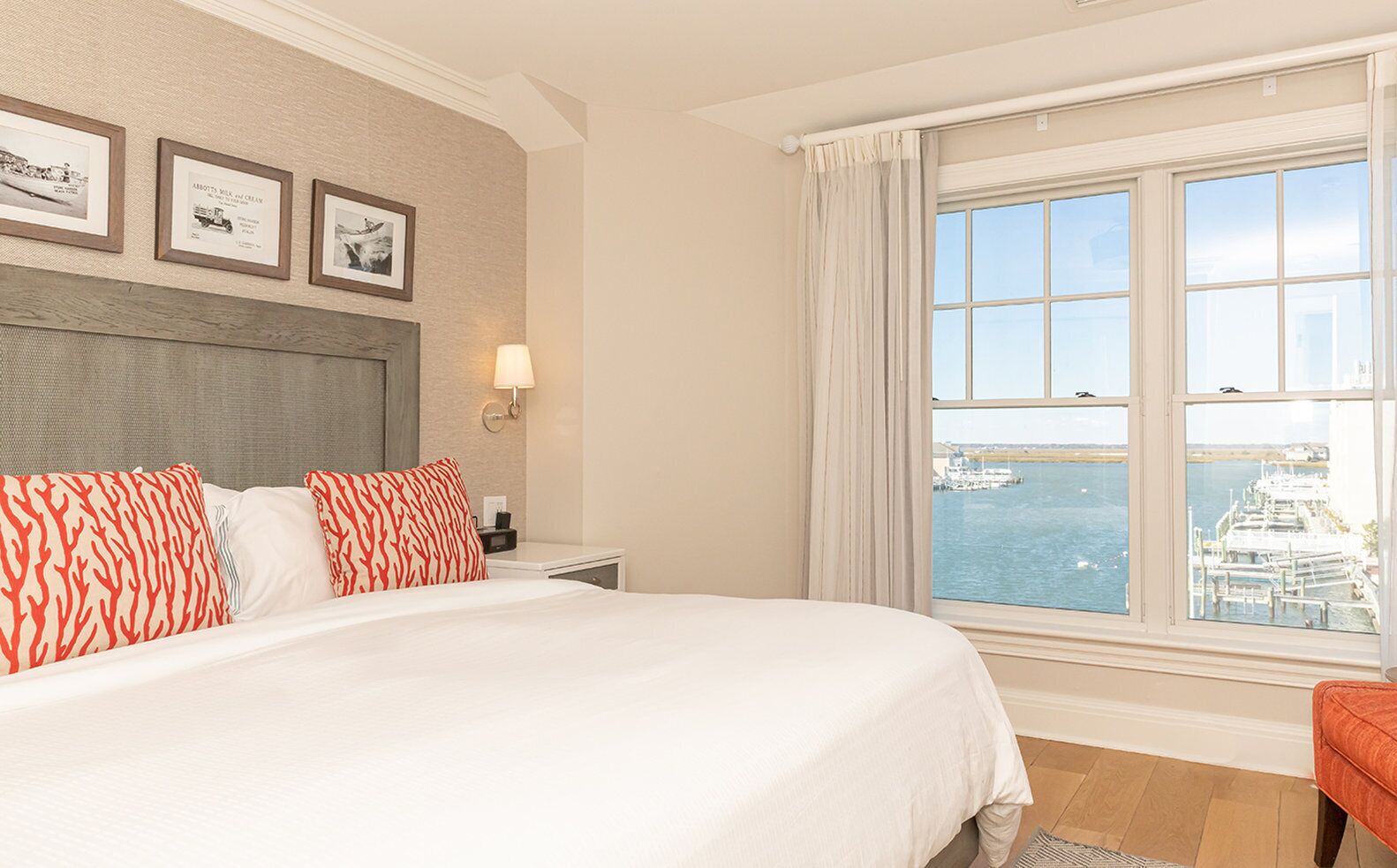 A hotel room with a large bed, a nightstand, a lamp, and a window with a view of the sea and boats.
