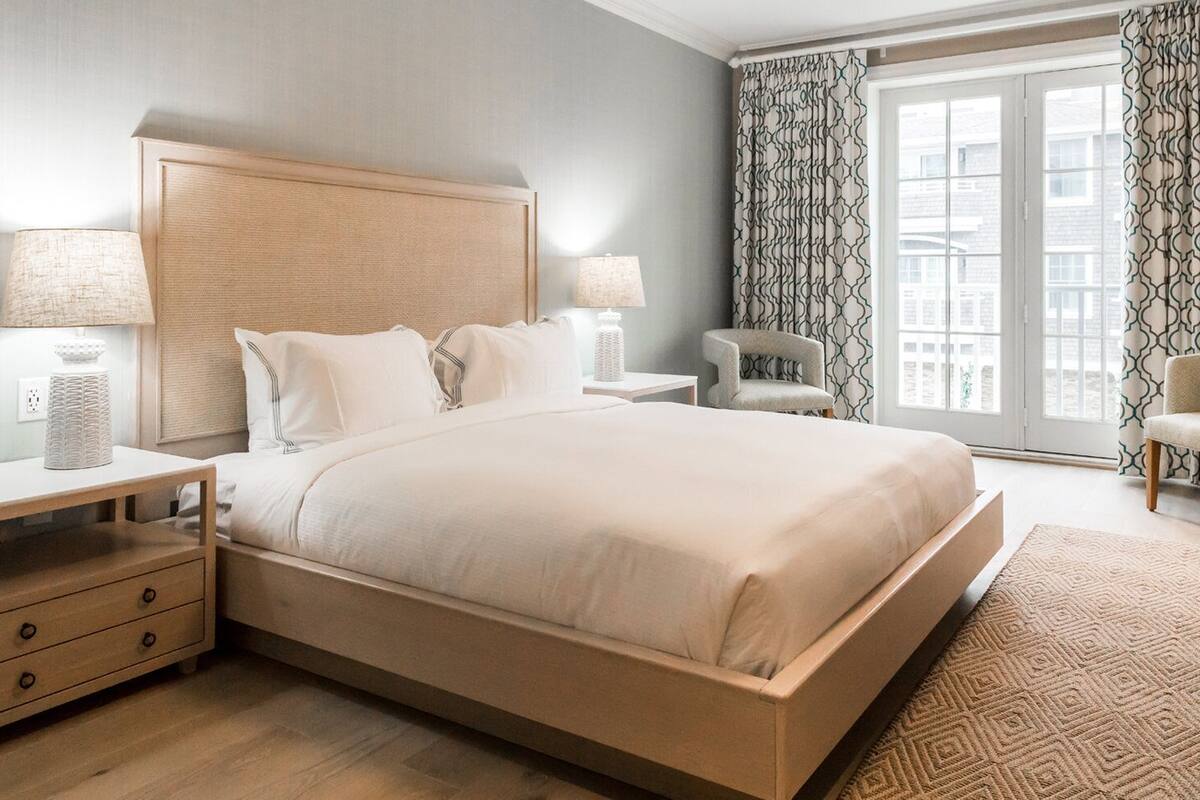 A modern hotel room with a large bed, two bedside tables, a chair, and a view of the city through the window.