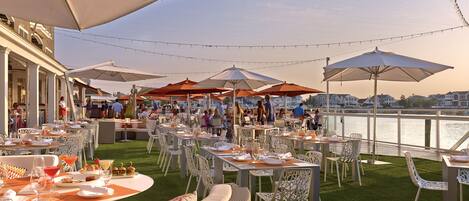 An outdoor dining area with tables and chairs, umbrellas, and a view of the water.