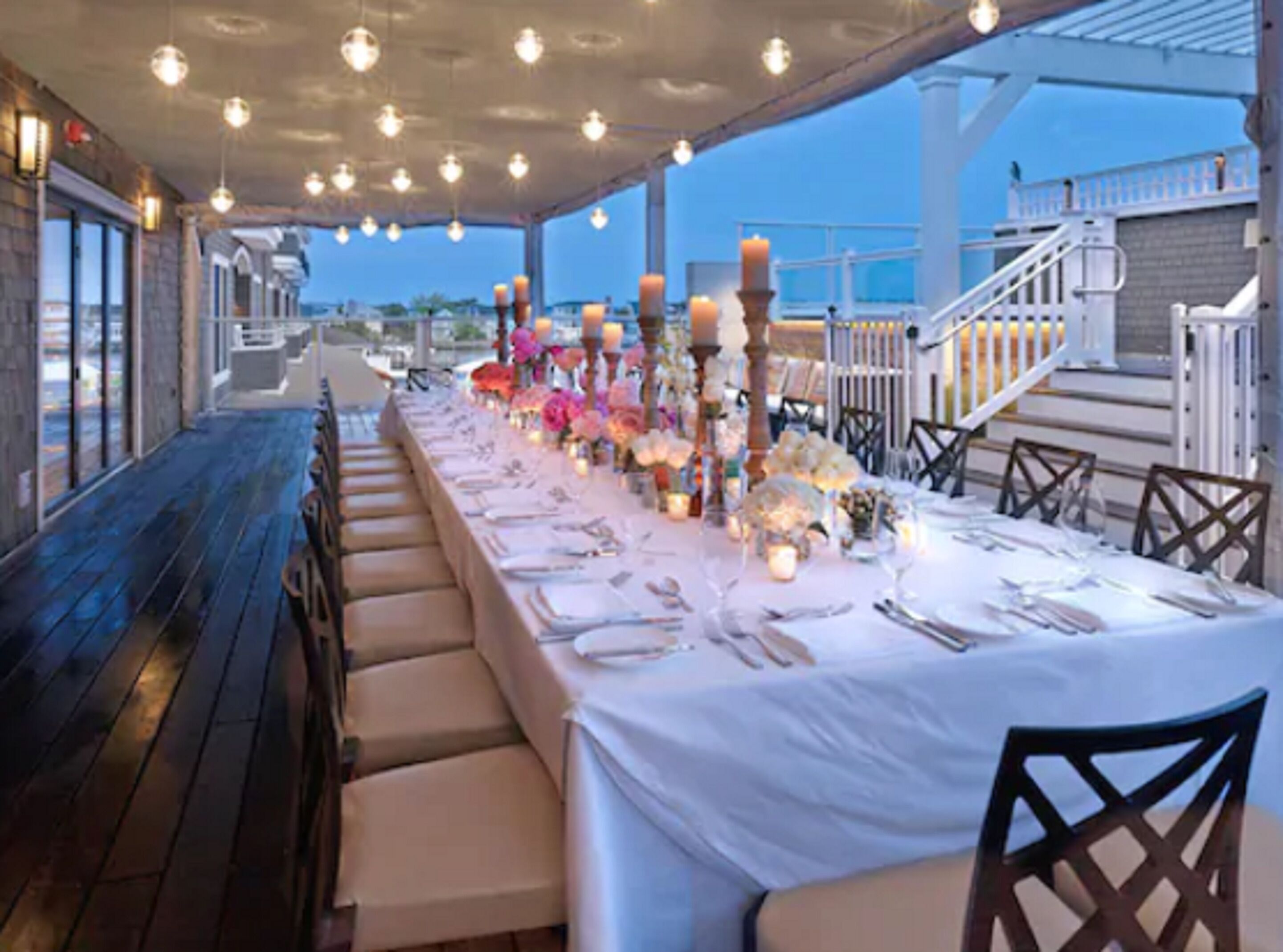 An outdoor dining area with a long table set for a meal, surrounded by chairs, and decorated with flowers and candles.