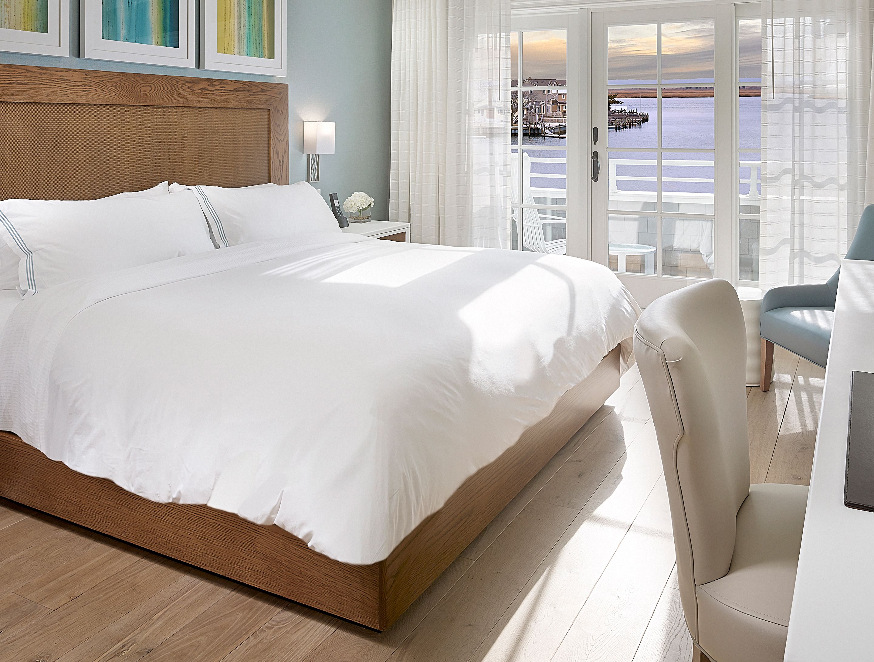 Habitación de hotel con una cama grande, cabecera de madera, mesita de noche con lámpara y vista al agua a través de puertas de cristal.