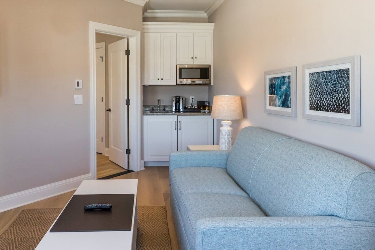 A living room with a light blue sofa, a remote control on a table, a lamp, and a kitchen in the background.