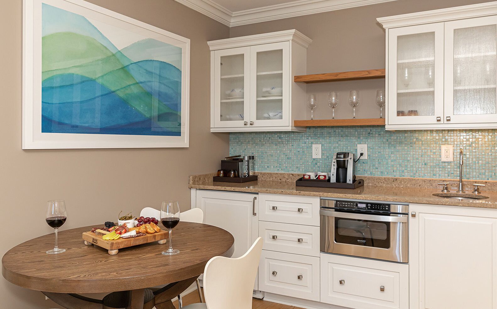 A modern kitchen with white cabinetry, a dining table with chairs, a coffee maker, and a large abstract painting on the wall.