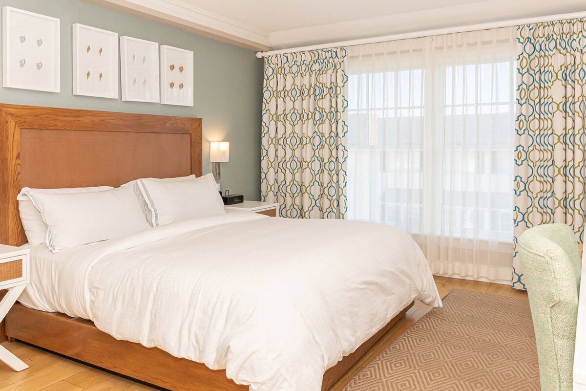 A neatly made bed in a hotel room with a wooden headboard, a bedside table with a lamp, and a patterned curtain.