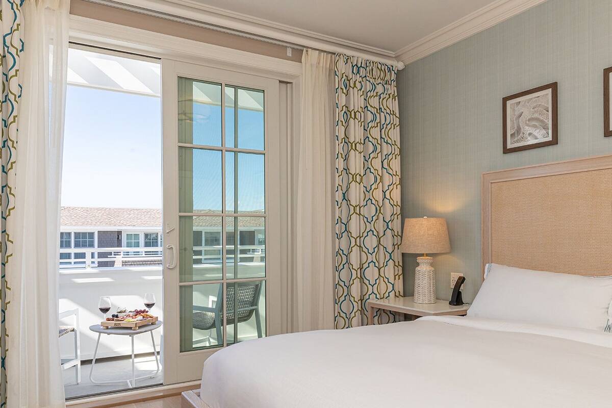A hotel room with a large bed, a nightstand with a lamp, a chair, and a balcony with a table and wine glasses.
