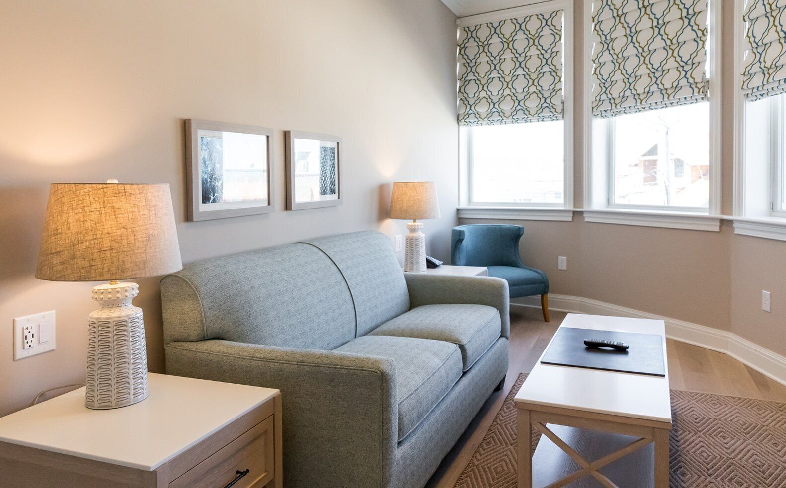 A living room with a grey sofa, two side tables with lamps, a blue armchair, and patterned window valances.