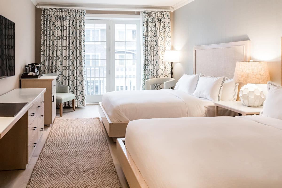 A hotel room with two beds, a desk, a chair, a lamp, and a view of the outside.