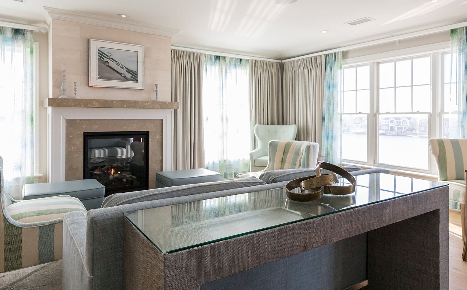 A modern living room with a glass coffee table, striped armchairs, a fireplace, and large windows with curtains.