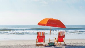 Two beach chairs with a red umbrella labeled "The Reeds" on a sandy beach with the ocean in the background. - The Reeds at Shelter Haven (Stone Harbor)