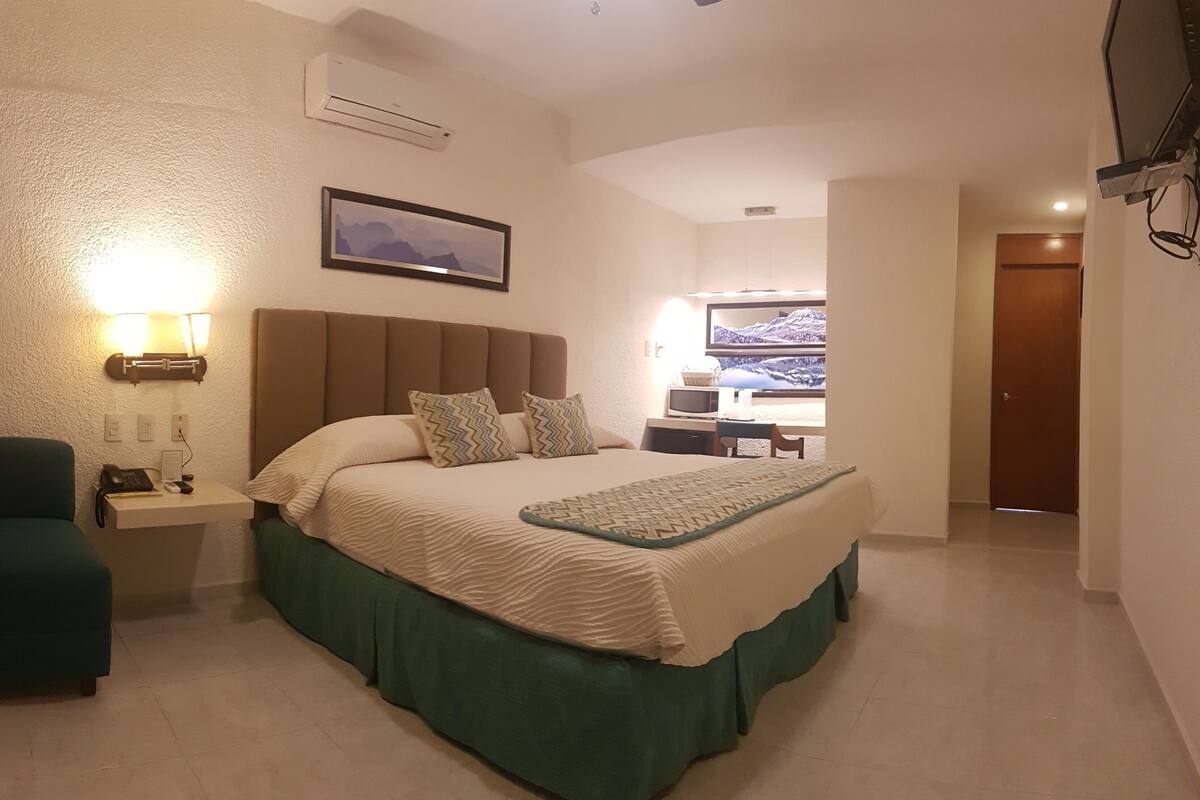 A hotel room with a bed, bedside table with a phone, lamp, and a chair. There is a desk with a computer and a TV in the background.