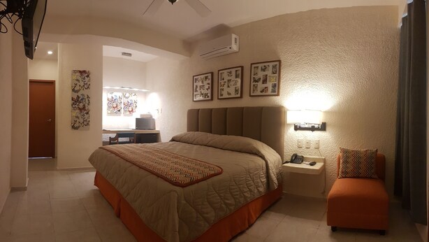 A hotel room with a large bed, a desk with a chair, a desk lamp, a telephone, and a small orange ottoman. - Hotel & Suites Nader (Cancun)