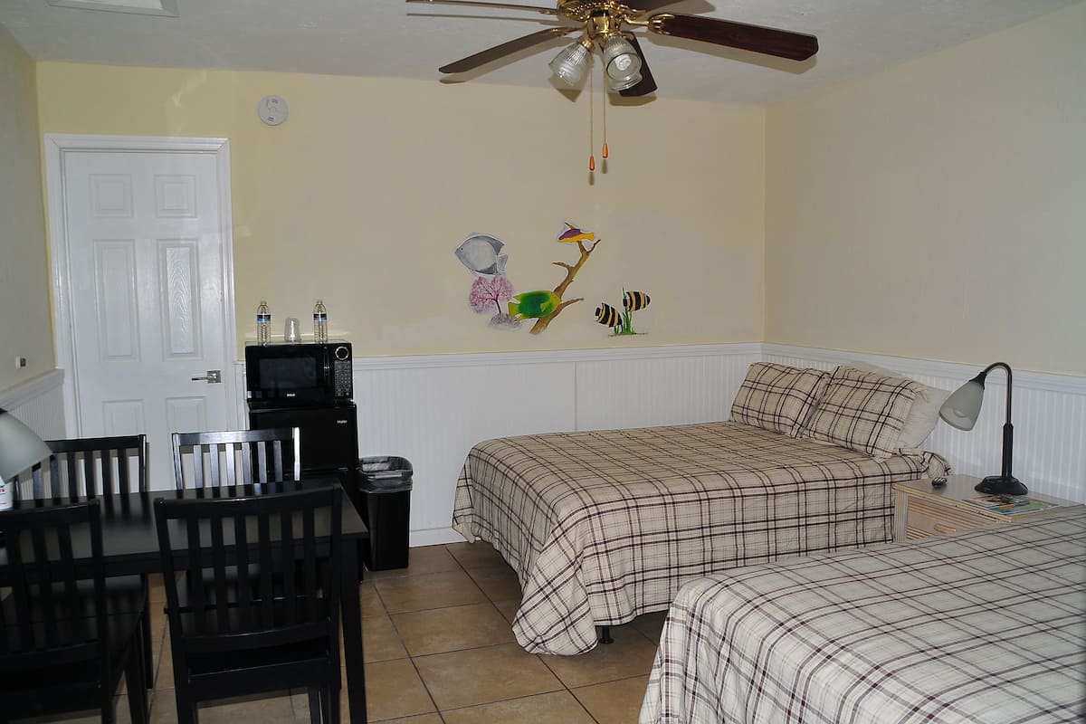 Une chambre avec deux lits, une table à manger avec des chaises, un ventilateur de plafond, un four à micro-ondes et une horloge murale.