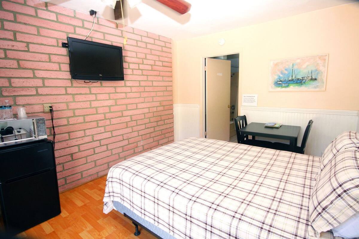 Une chambre avec un mur en briques, un téléviseur à écran plat fixé au-dessus d’une commode, un lit recouvert d’une couverture à carreaux, une petite table avec une chaise et un tableau accroché au mur.