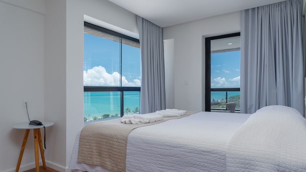 A modern bedroom with a large bed, a small side table, and a view of the ocean through large windows. - Vistamar Hotel (Maceió)