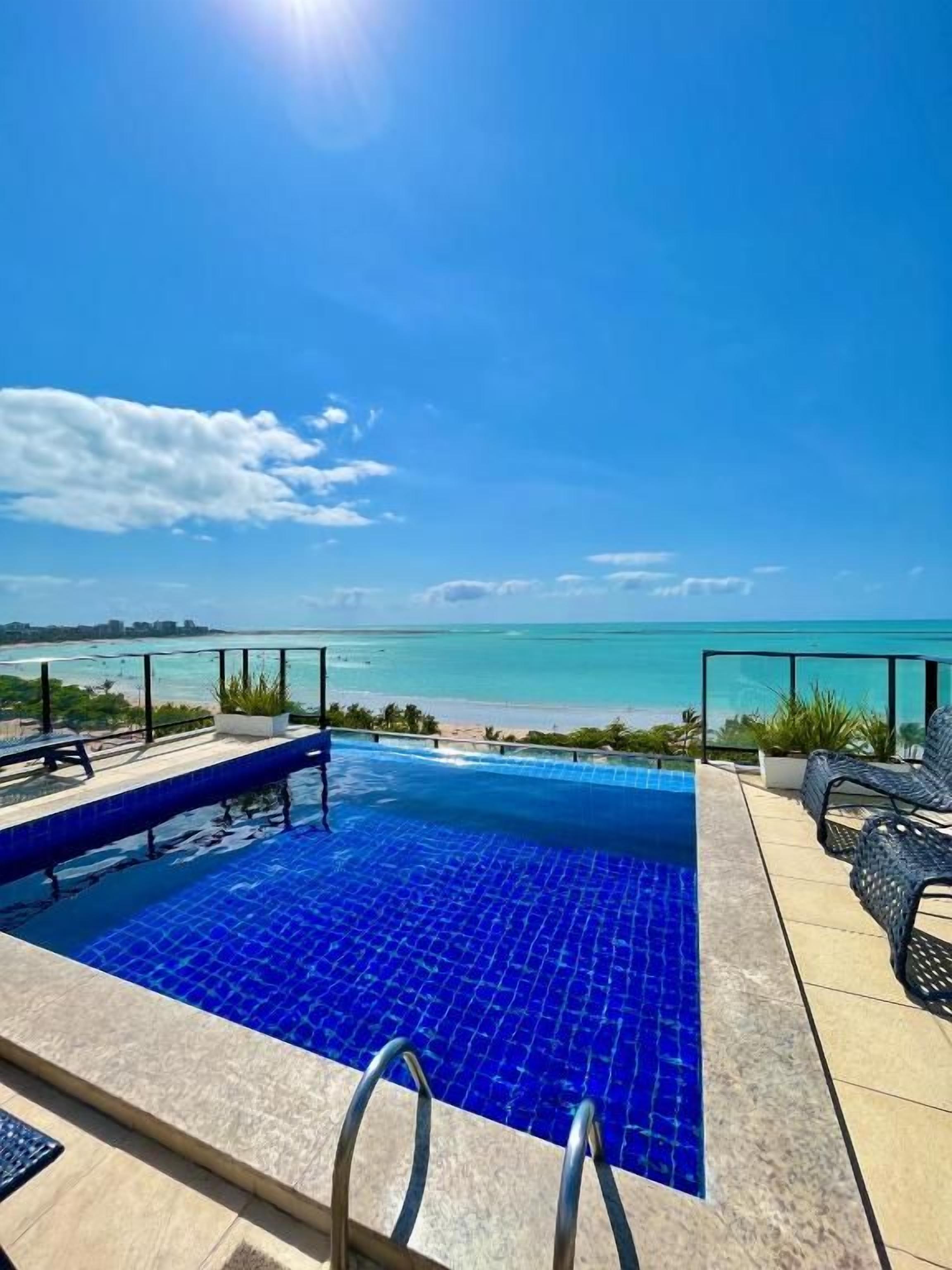 Una piscina con vista a la playa, rodeada de sillones de descanso y un cielo despejado.