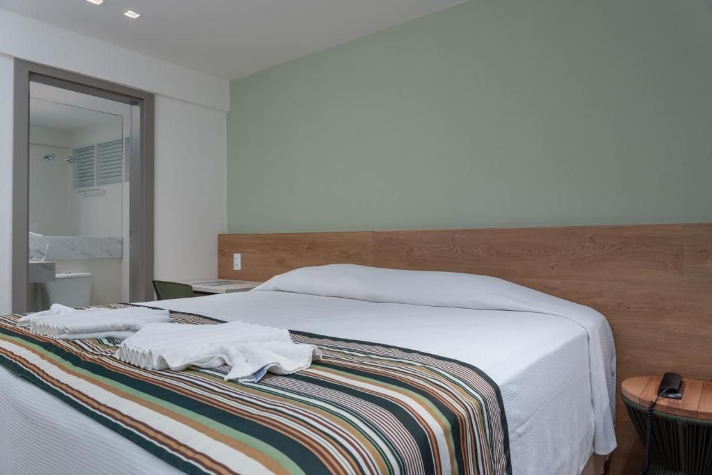Habitación de hotel con cama, cabecera de madera, mesita de noche con lámpara y baño visible a través de una puerta abierta.