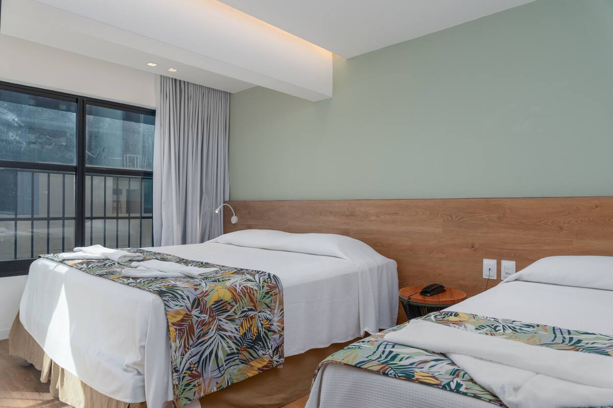 A hotel room with two beds, a wooden headboard, a large window with curtains, and a bedside table with a phone.