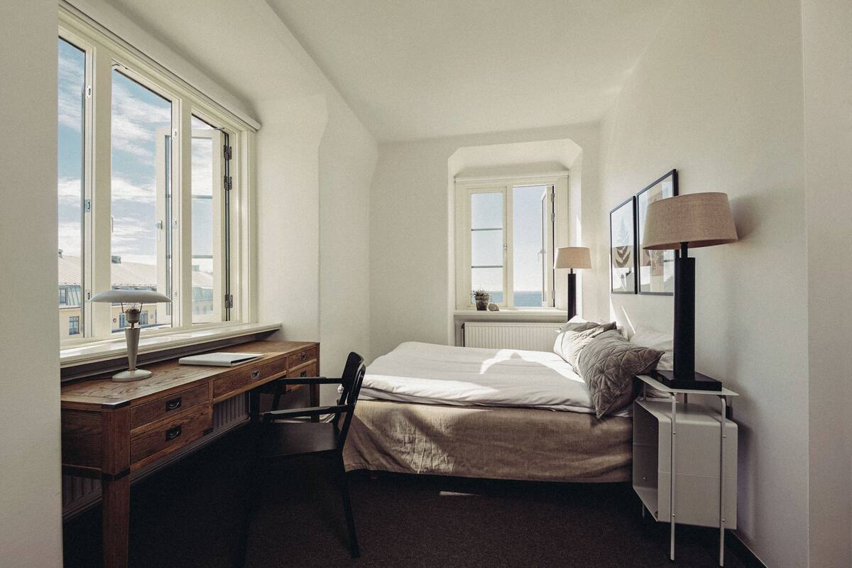 Ein Schlafzimmer mit Bett, Schreibtisch und Stuhl. Zwei Fenster sorgen für natürliches Licht.