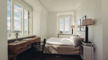 A bedroom with a bed, a desk, and a chair. There are two windows providing natural light.