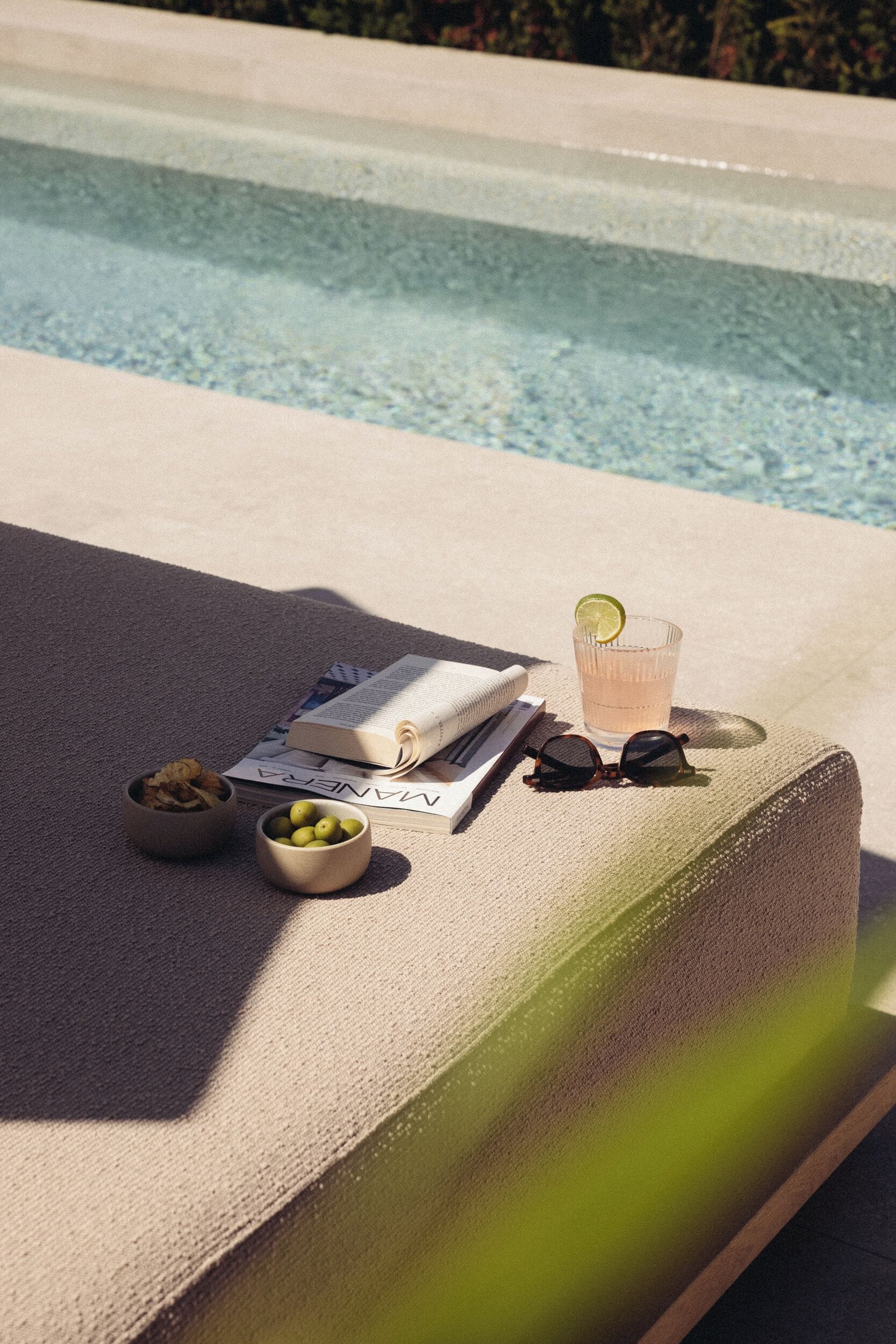 A poolside lounge area with a book, sunglasses, a glass of drink, and a bowl of lemons.