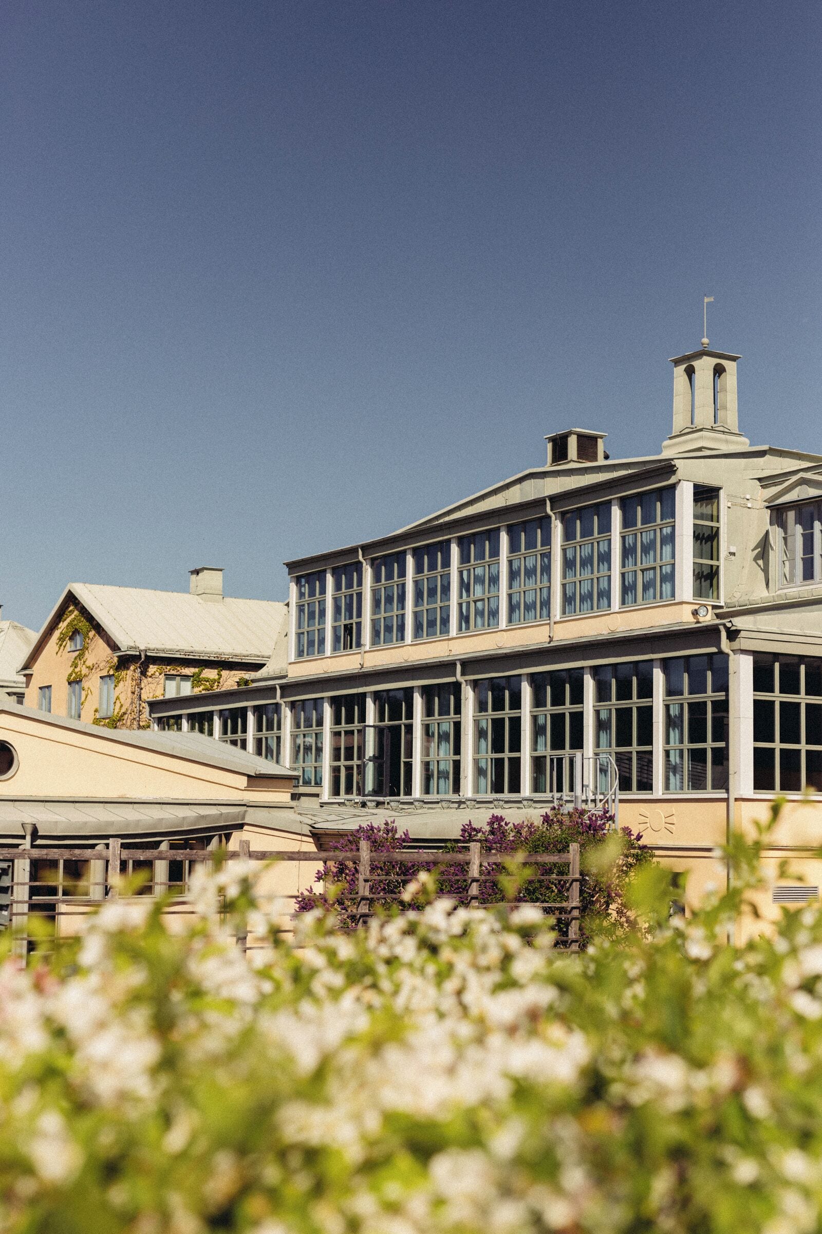 Un bâtiment doté d’une grande façade en verre, d’une tour horloge et d’un jardin où l’on peut voir, au premier plan, des fleurs blanches.