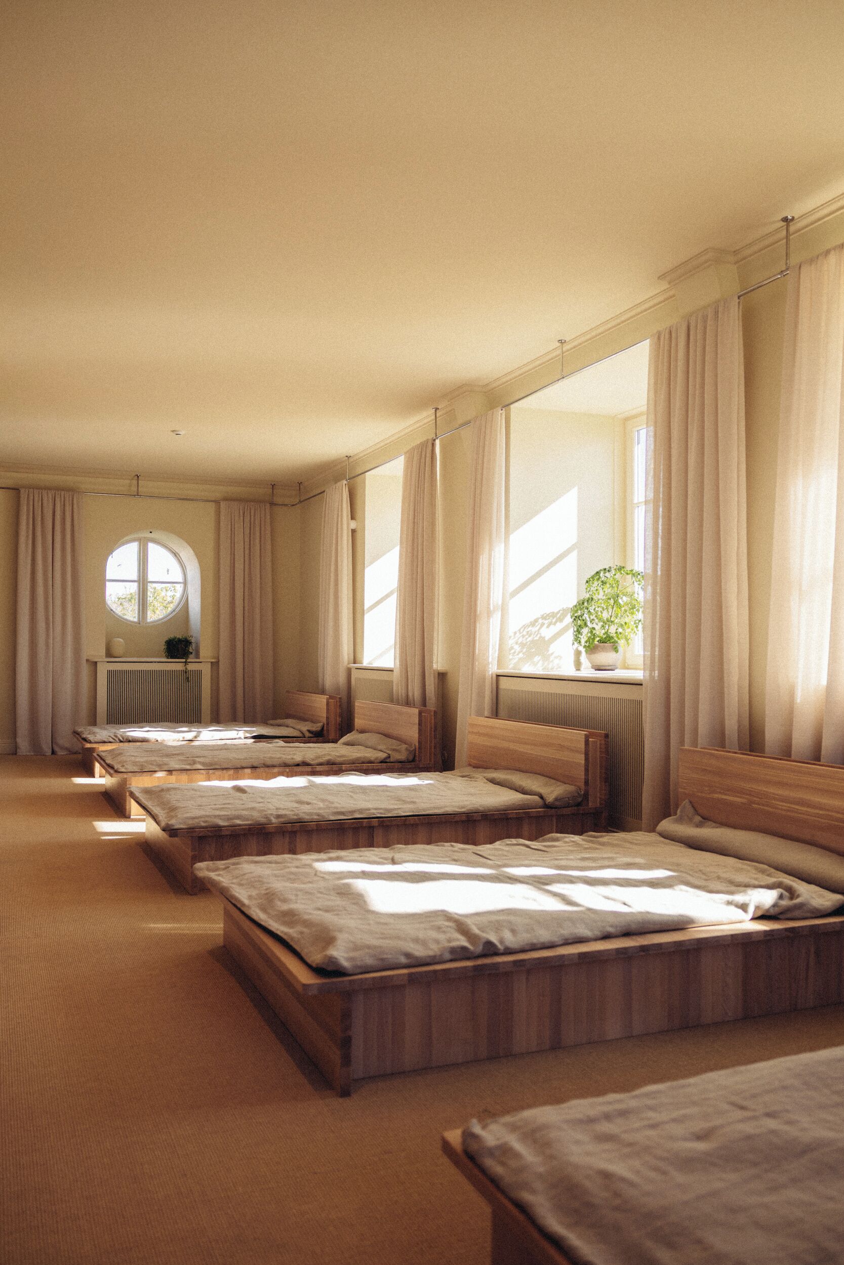 A hotel room with four single beds, a round window, a radiator, and a potted plant by the window.