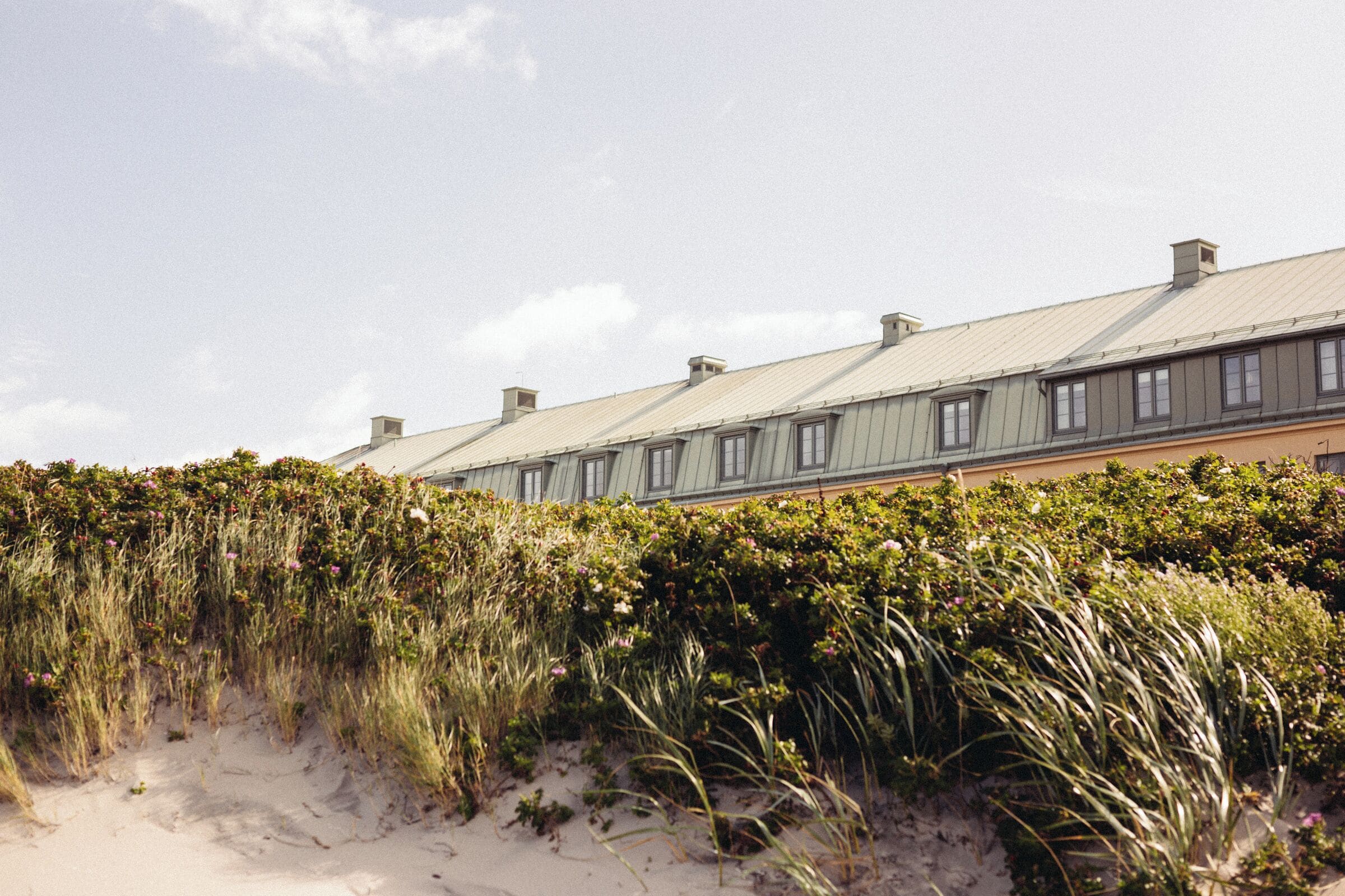 A row of buildings with a flat roof, surrounded by dense vegetation and sand dunes.