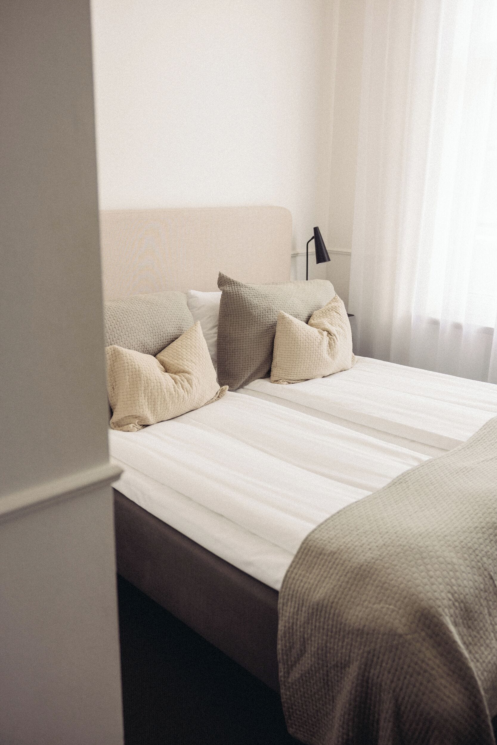 Una cama bien hecha con un cabecero color beige y almohadas, una lámpara de noche y vistas a una ventana con cortinas.
