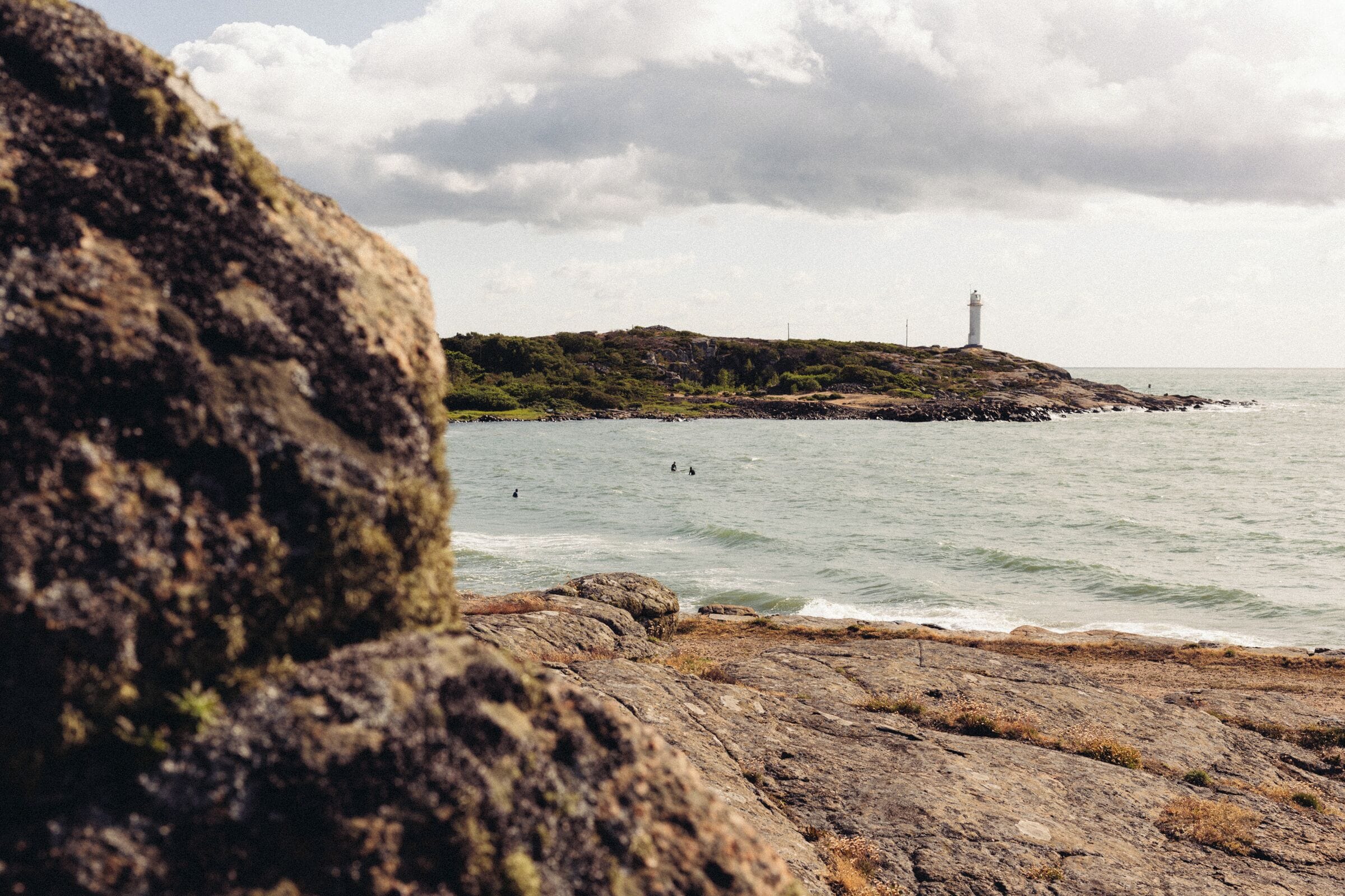 A rocky shoreline with a lighthouse in the distance and a few people in the water.
