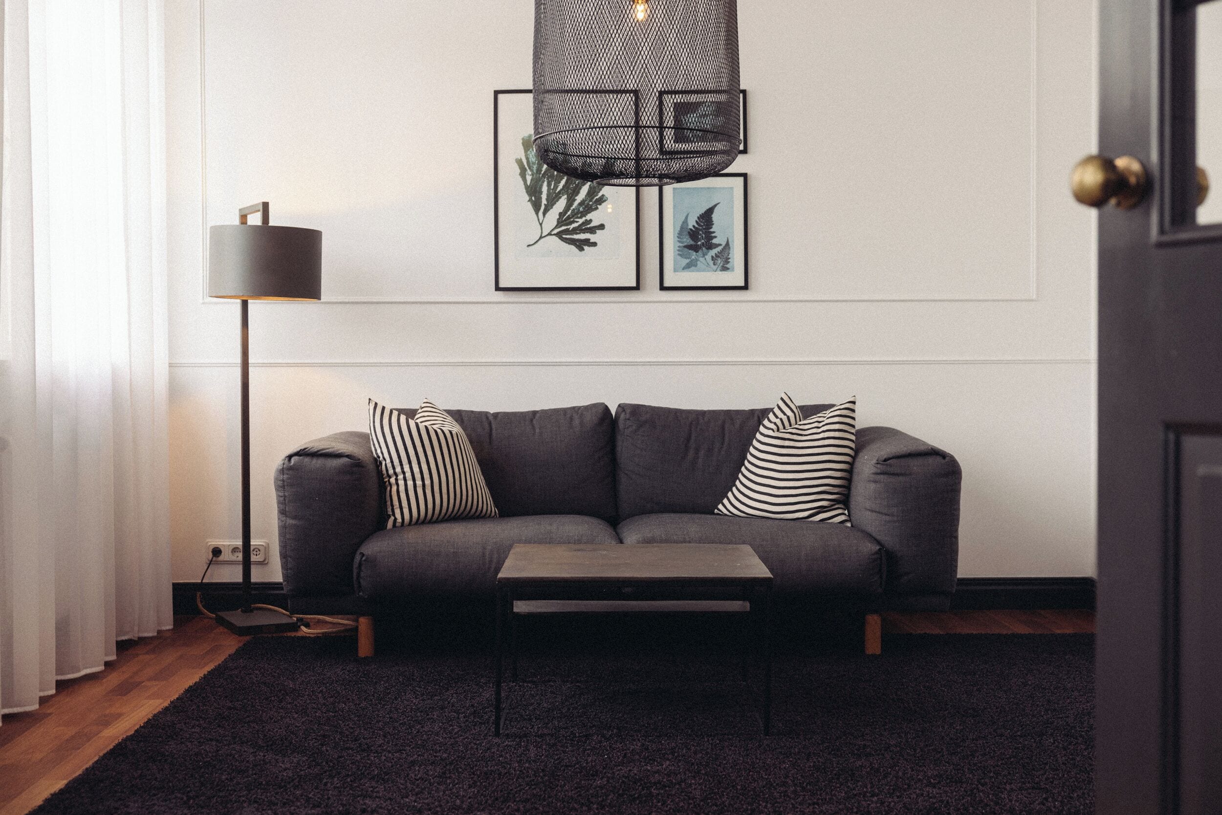 Un salon moderne avec un canapé gris, des coussins rayés noirs et blancs, une table basse sombre, un lampadaire et des reproductions botaniques encadrées accrochées au mur.