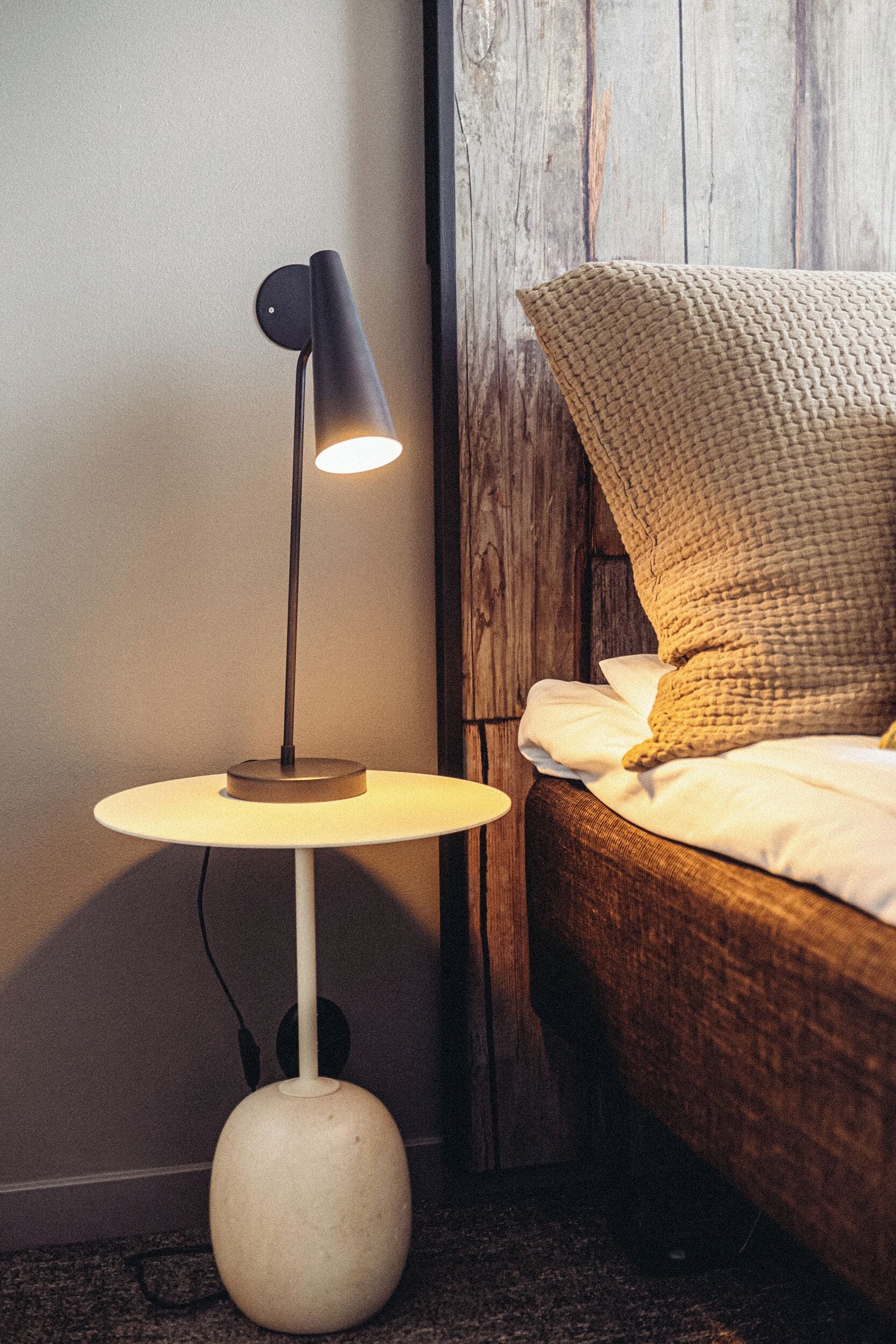 A bedside table with a lamp, a bed with a textured blanket, and a wooden wall.