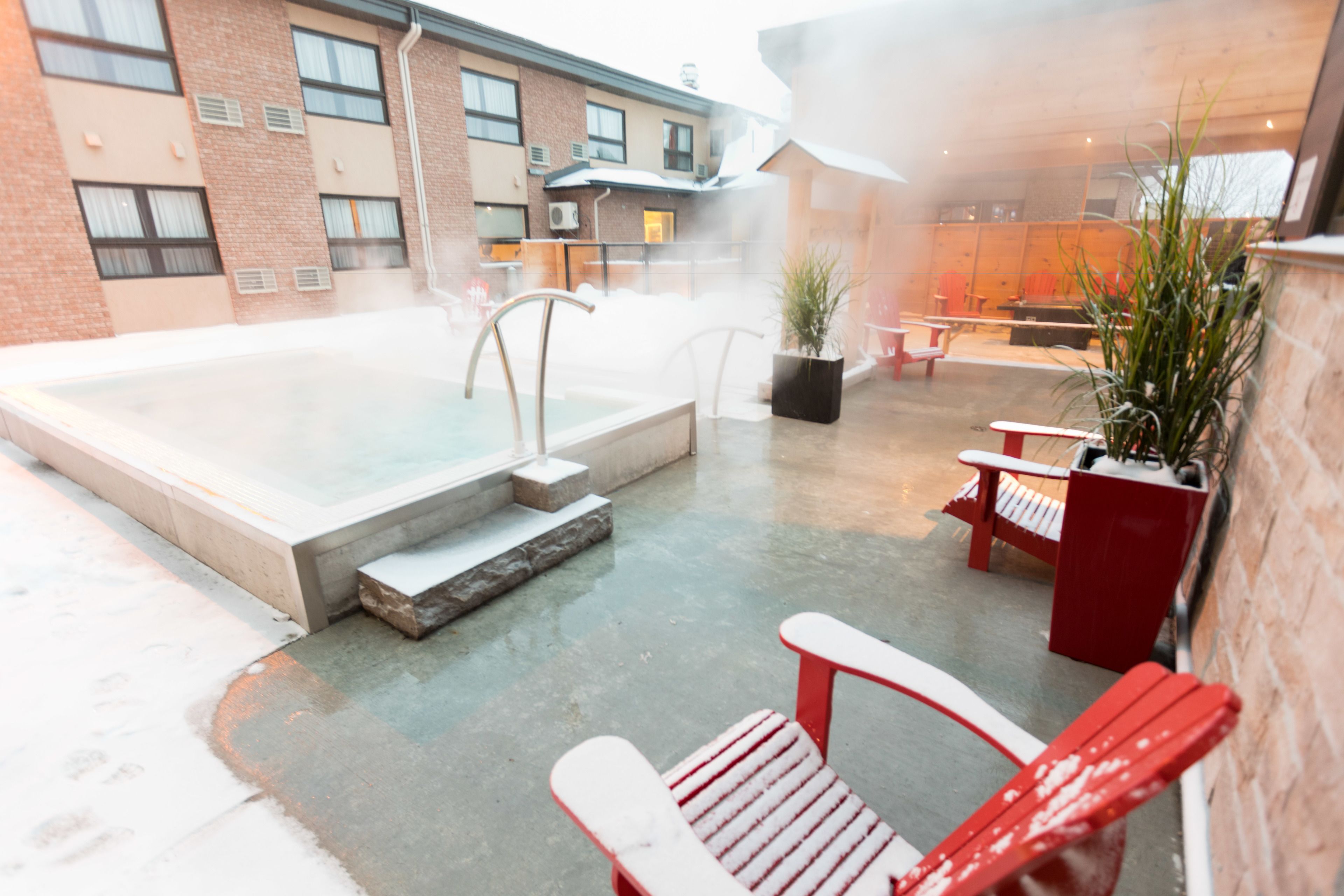 A heated outdoor pool area with red Adirondack chairs, a potted plant, and a brick building in the background.