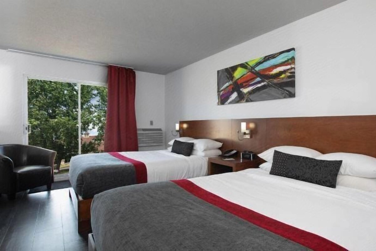 A hotel room with two beds, a chair, a painting, and a view of trees.