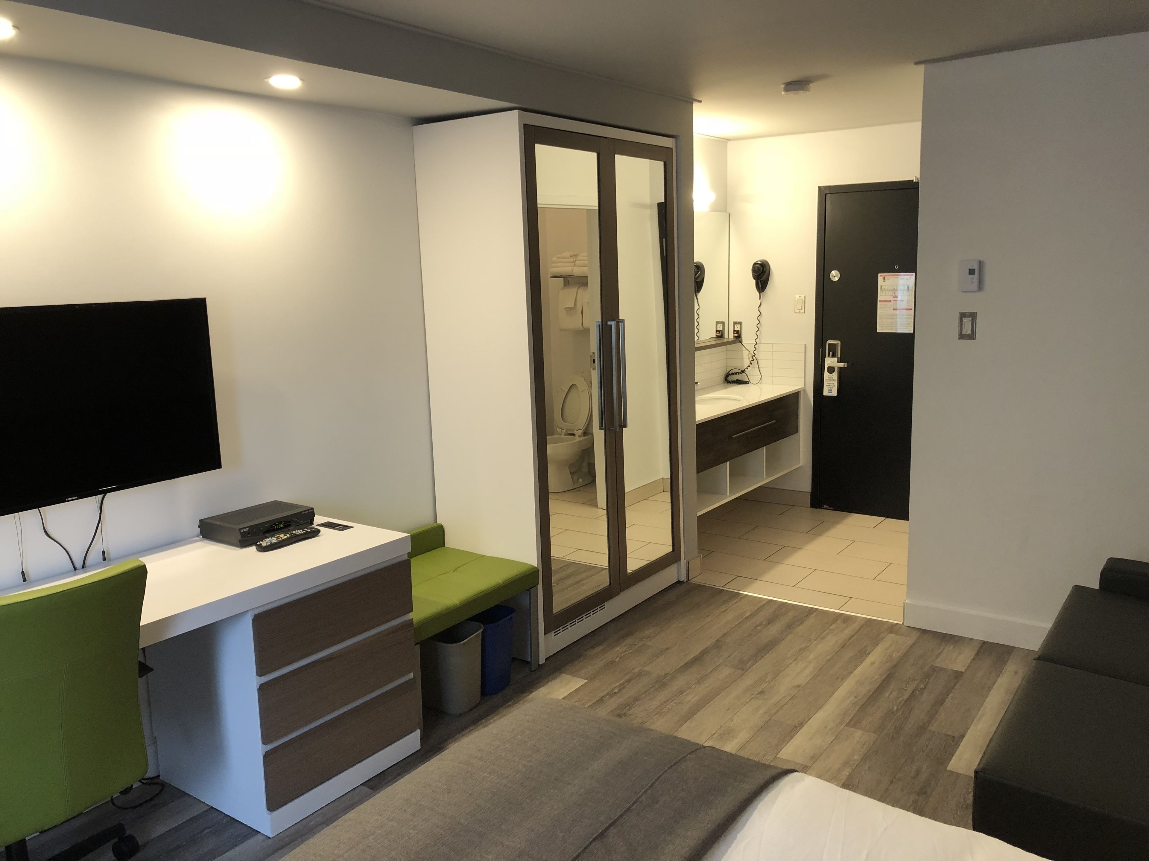 Una habitación de hotel moderna con televisión de pantalla plana, un escritorio con una silla, un espejo, un armario, un baño con inodoro y lavabo, y una puerta con cerradura.