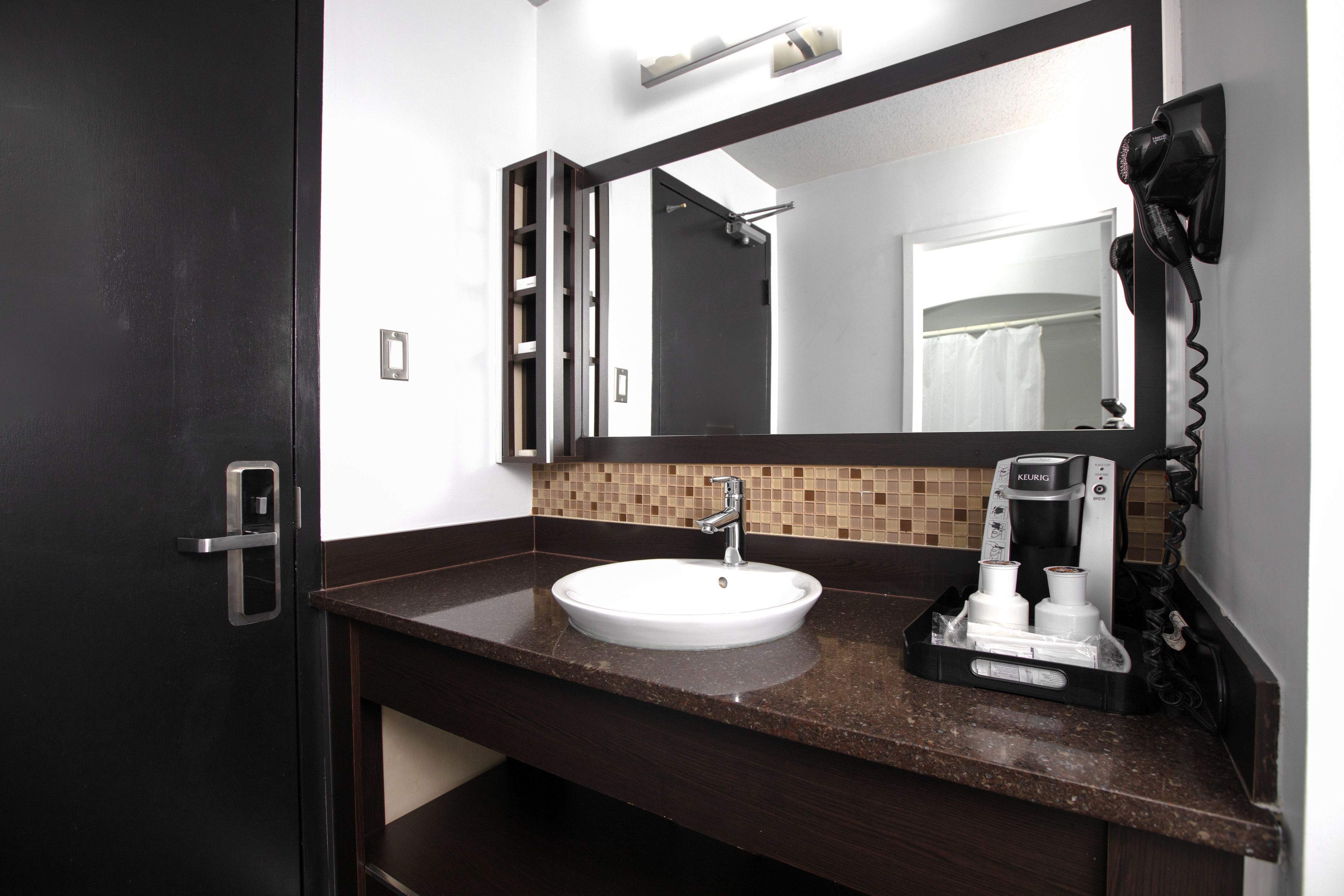 Un bagno con un piano d'appoggio marrone scuro, un lavandino bianco, un asciugacapelli nero e una macchina per il caffè.