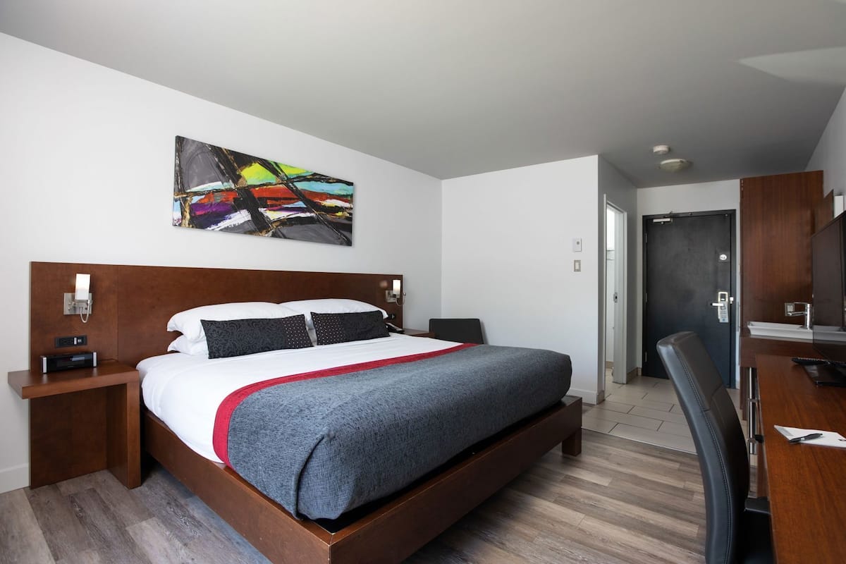 A hotel room with a bed, bedside tables, a desk, a chair, a painting on the wall, and a door.