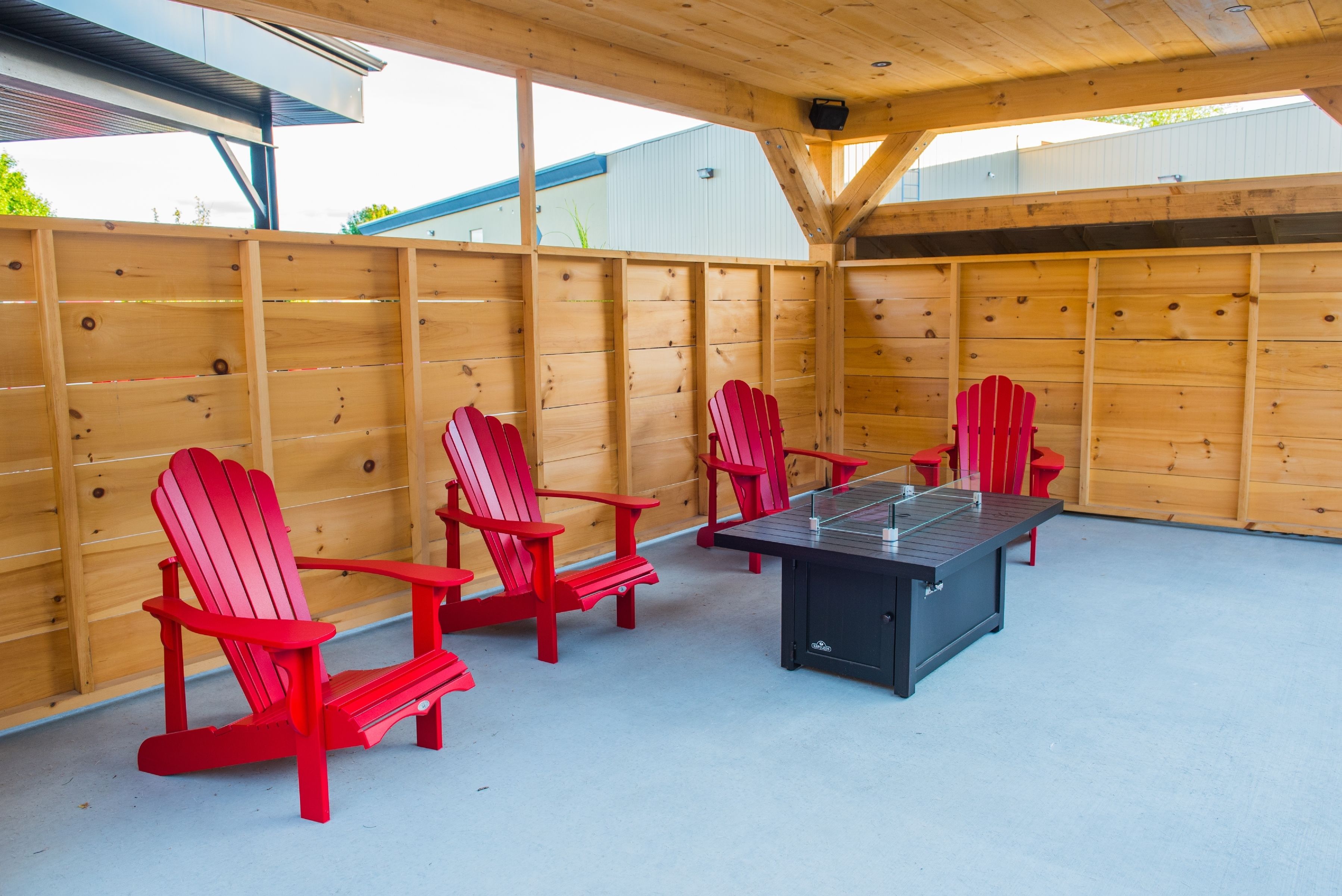An outdoor seating area with wooden walls, red Adirondack chairs, and a black table.