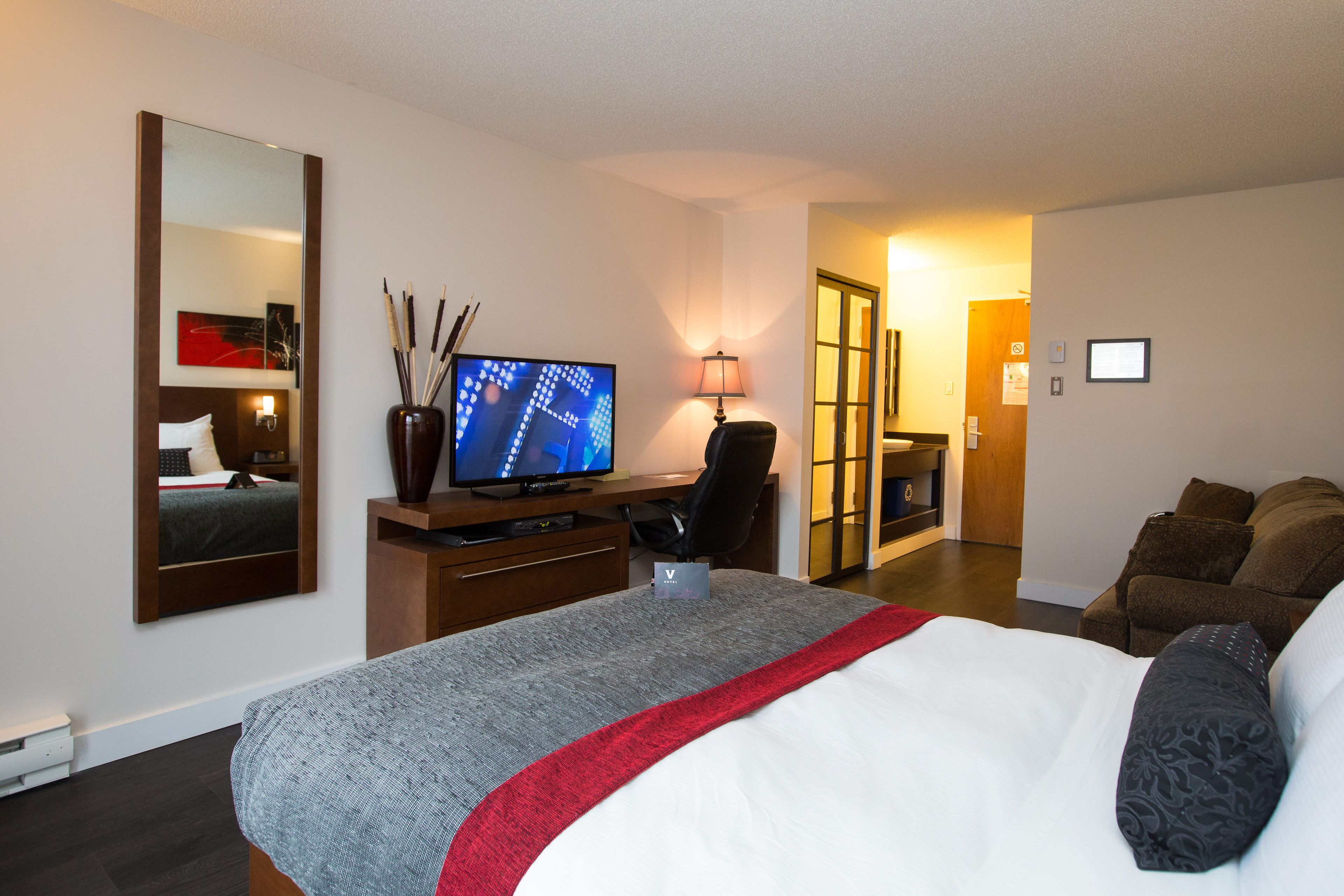 A hotel room with a bed, a desk with a computer, a TV, a mirror, a lamp, and a door leading to another room.