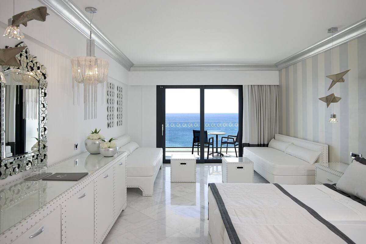 A modern hotel room with a large bed, a sitting area with a view of the ocean, a chandelier, and a decorative mirror.