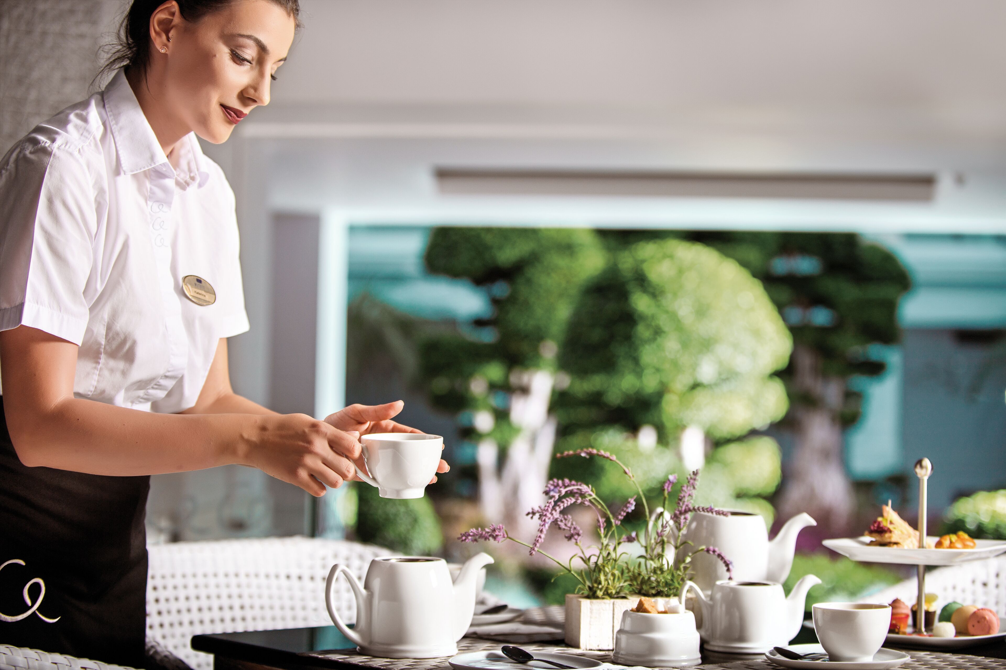 A person serving tea in a room with a table set for tea service, including a teapot, cups, and a tray with pastries.