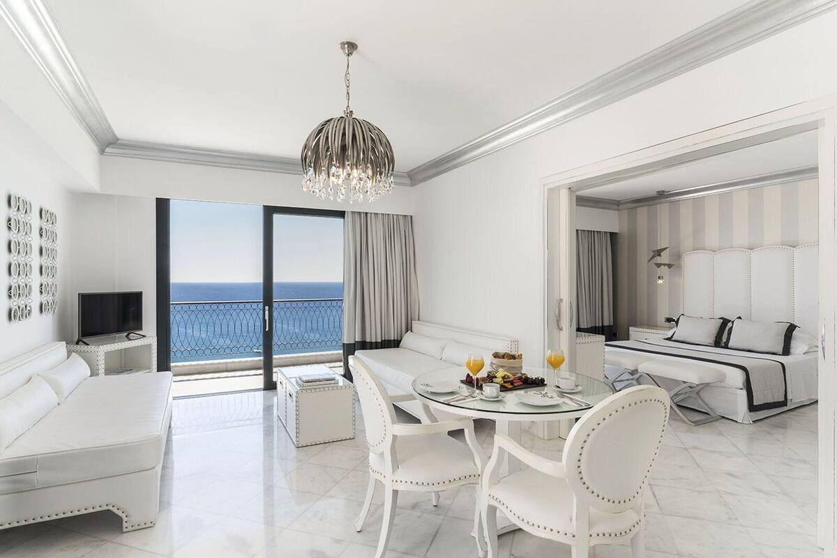 A modern hotel room with a balcony, a dining area with white furniture, and a large bed with a headboard.