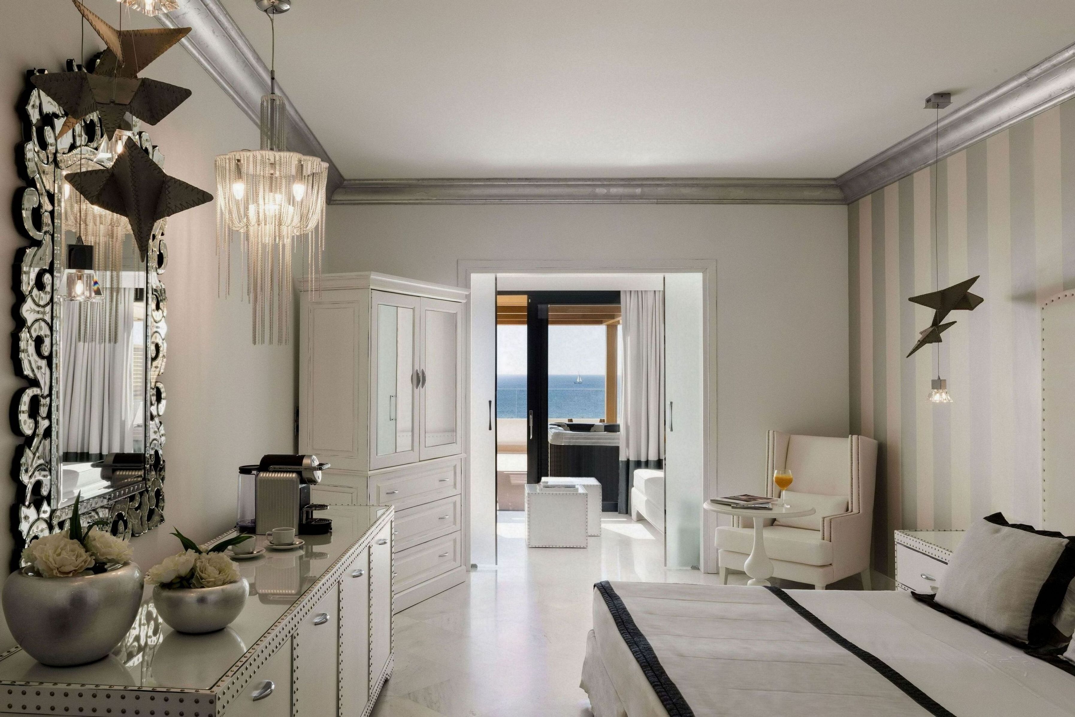 A modern hotel room with a large bed, a vanity with a mirror, a chandelier, and a view of the sea through an open door.