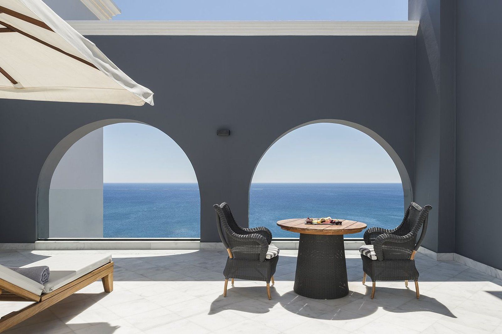 Un balcón con dos sillas de mimbre y una mesa redonda, con vista a un mar azul y despejado.