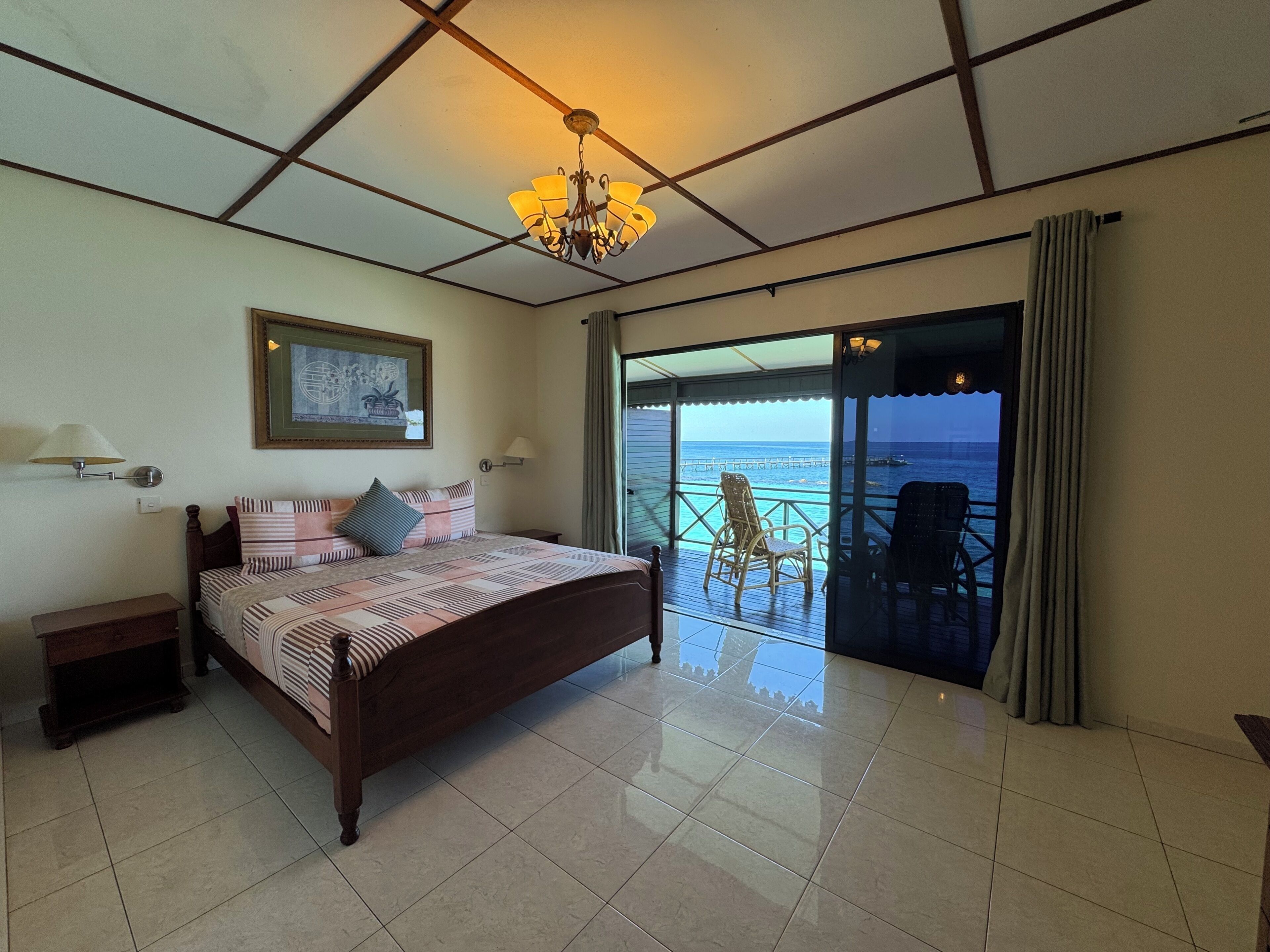 Un dormitorio con cama, candelabro, cuadro enmarco, mesita de noche, ventana con vista al océano y balcón con sillas.