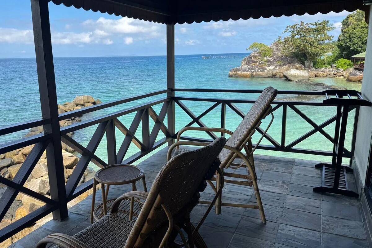 A covered outdoor seating area with wicker chairs and a small round table, overlooking a turquoise sea and rocky coastline.