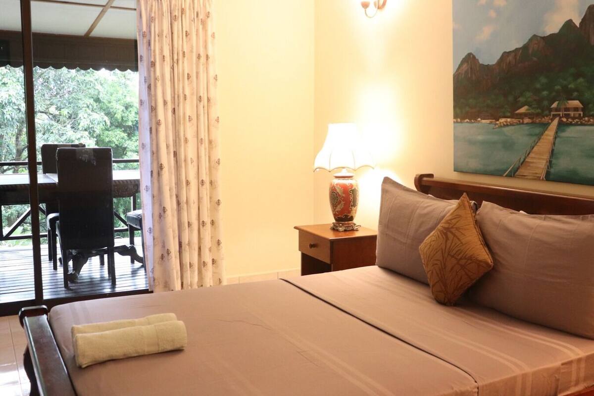 A hotel room with a bed, a nightstand with a lamp, a painting on the wall, and a view of a deck and greenery outside.