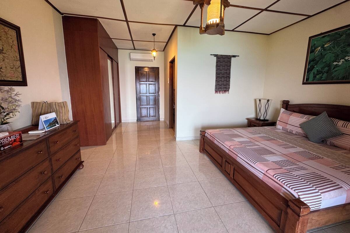 A bedroom with a large bed, a wooden dresser, a desk with a lamp, a ceiling fan, and a door leading to another room.